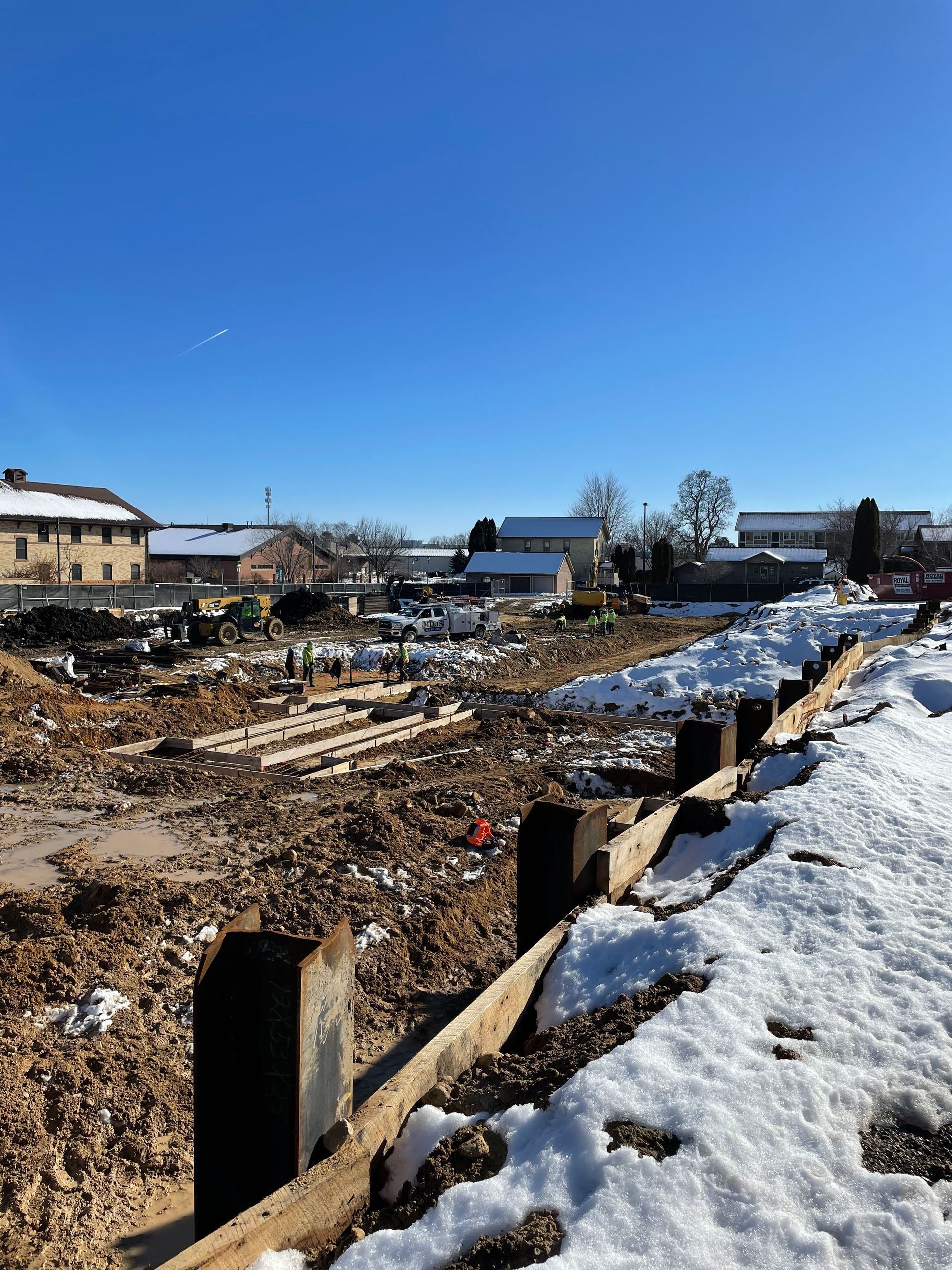 Construction site with dirt, snow, and wooden barriers under a blue sky, with buildings in the background.