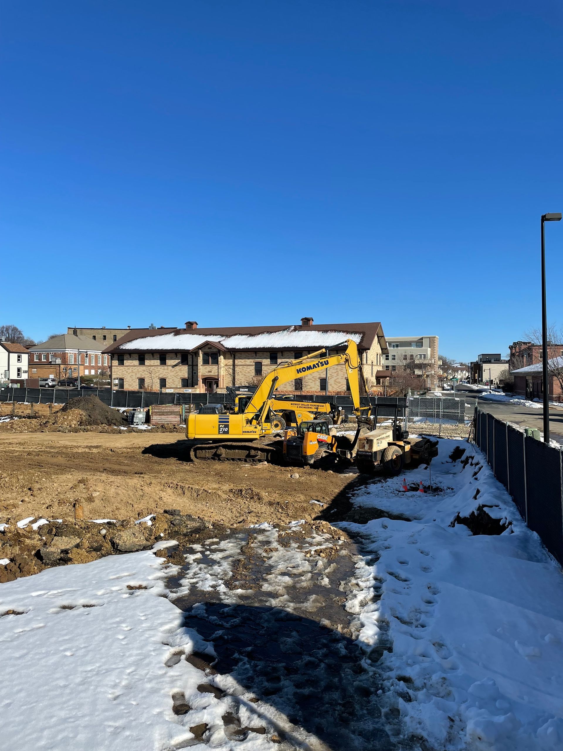 Yellow excavator on a construction site with brown soil and patches of snow; buildings in the background.