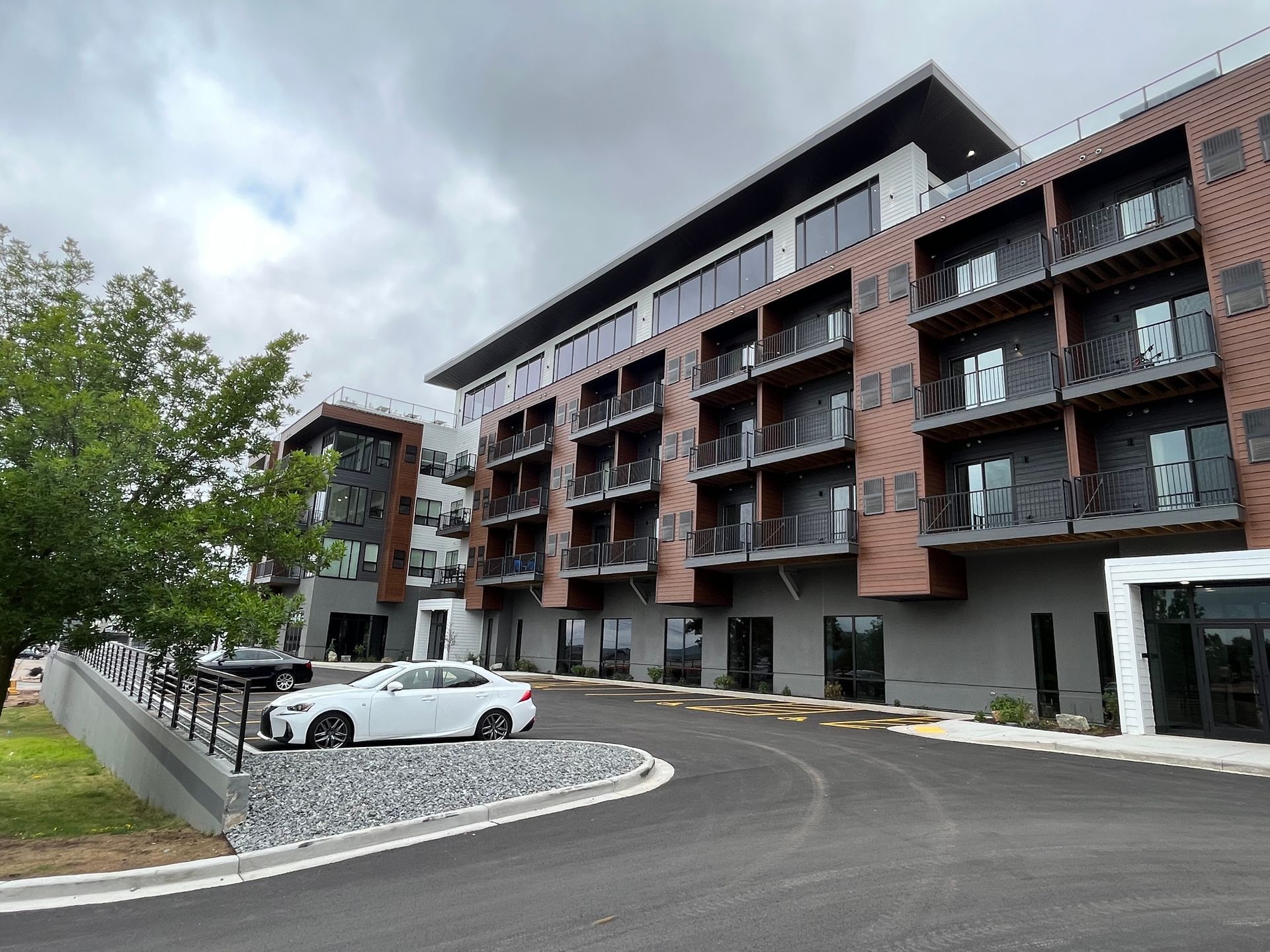 Modern multi-story building with brown and gray facade, balconies, and a parked white car. Overcast sky.