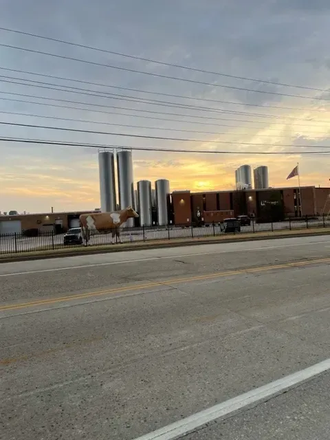 A factory with several tall silver silos at sunset; brown brick building, cars, and utility lines in the foreground.
