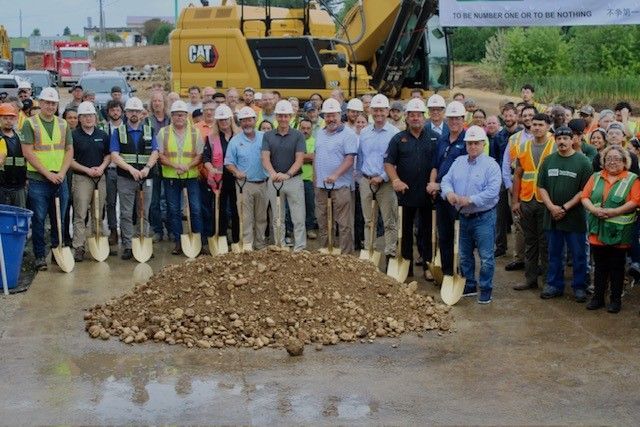 Groundbreaking ceremony with a group holding shovels over a pile of dirt. A large CAT machine is in the background.