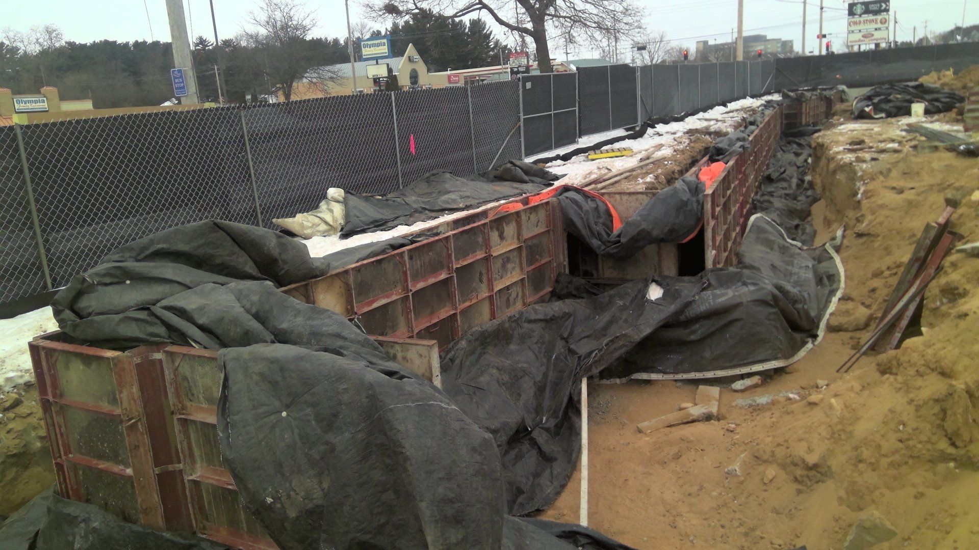 Construction site with a metal structure, dirt, and black tarp next to a black fence.