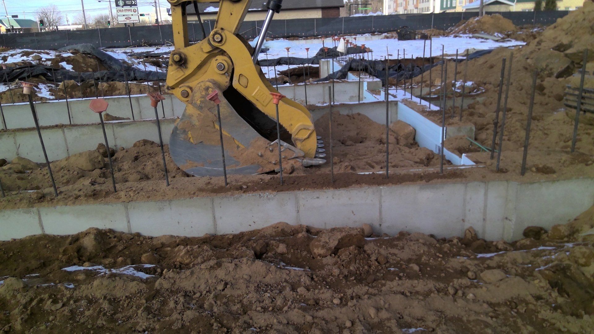 Yellow excavator scooping dirt from a construction site with exposed foundation walls and rebar.