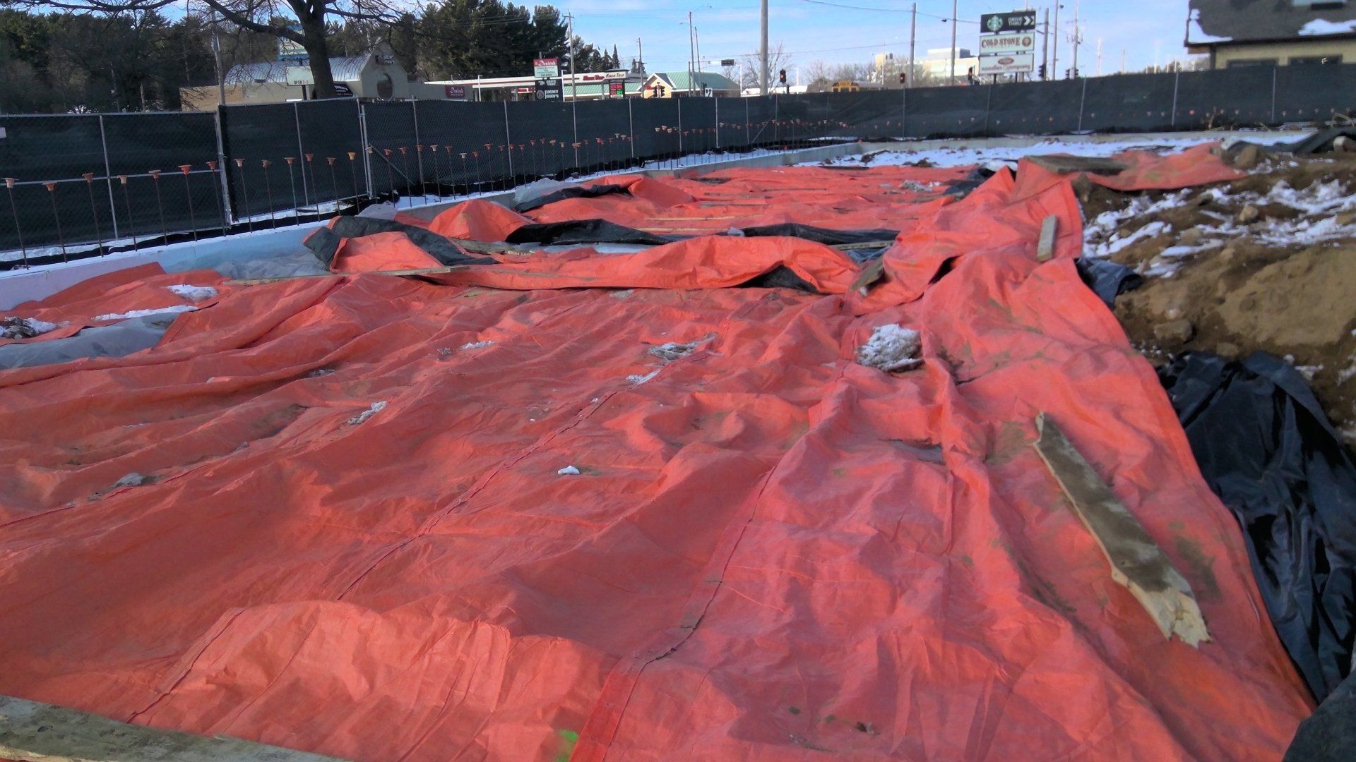 Orange tarp covering an excavation site with some snow and a black fence in the background.