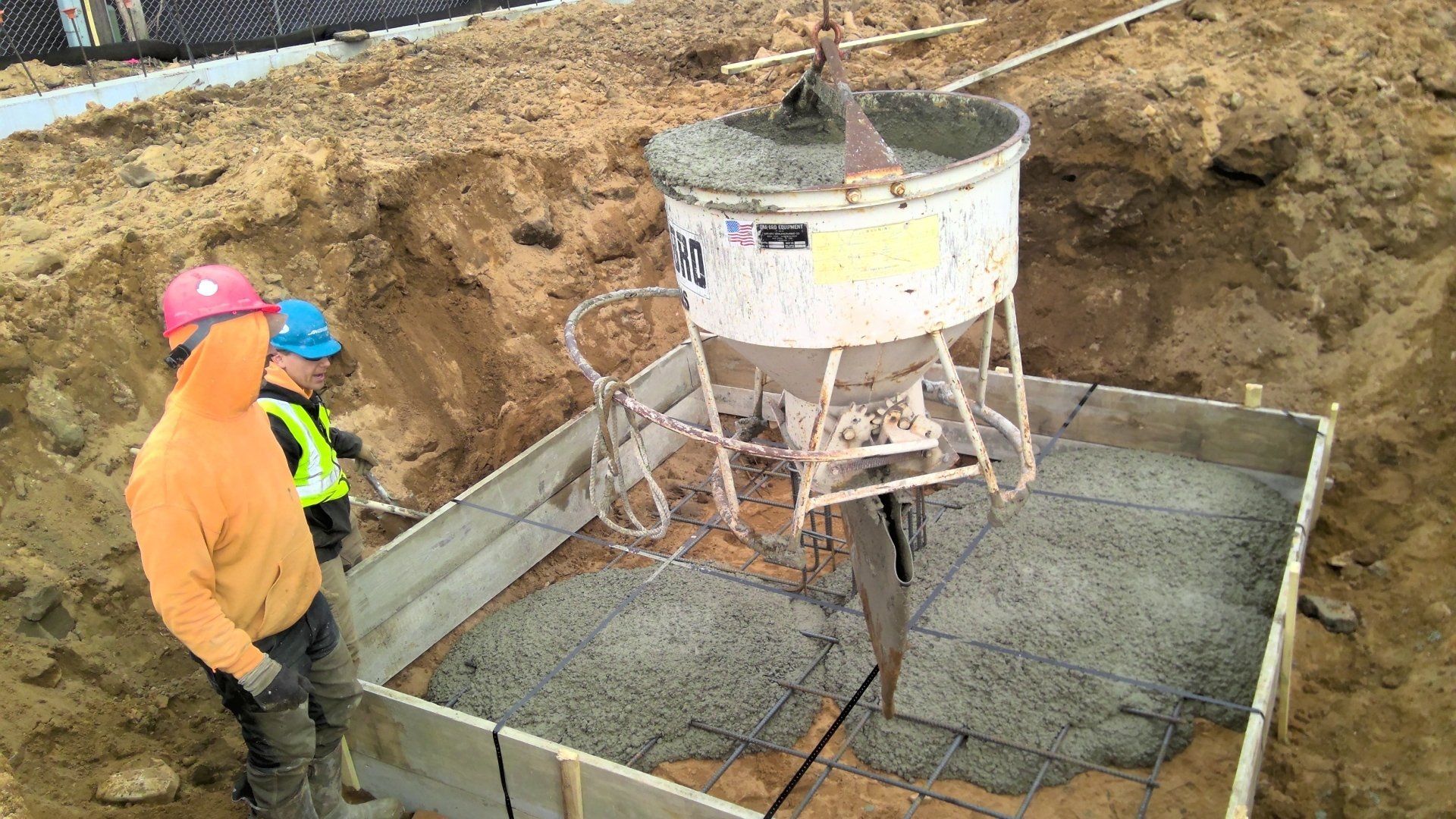 Workers pouring concrete into a wooden form, using a suspended container at a construction site.