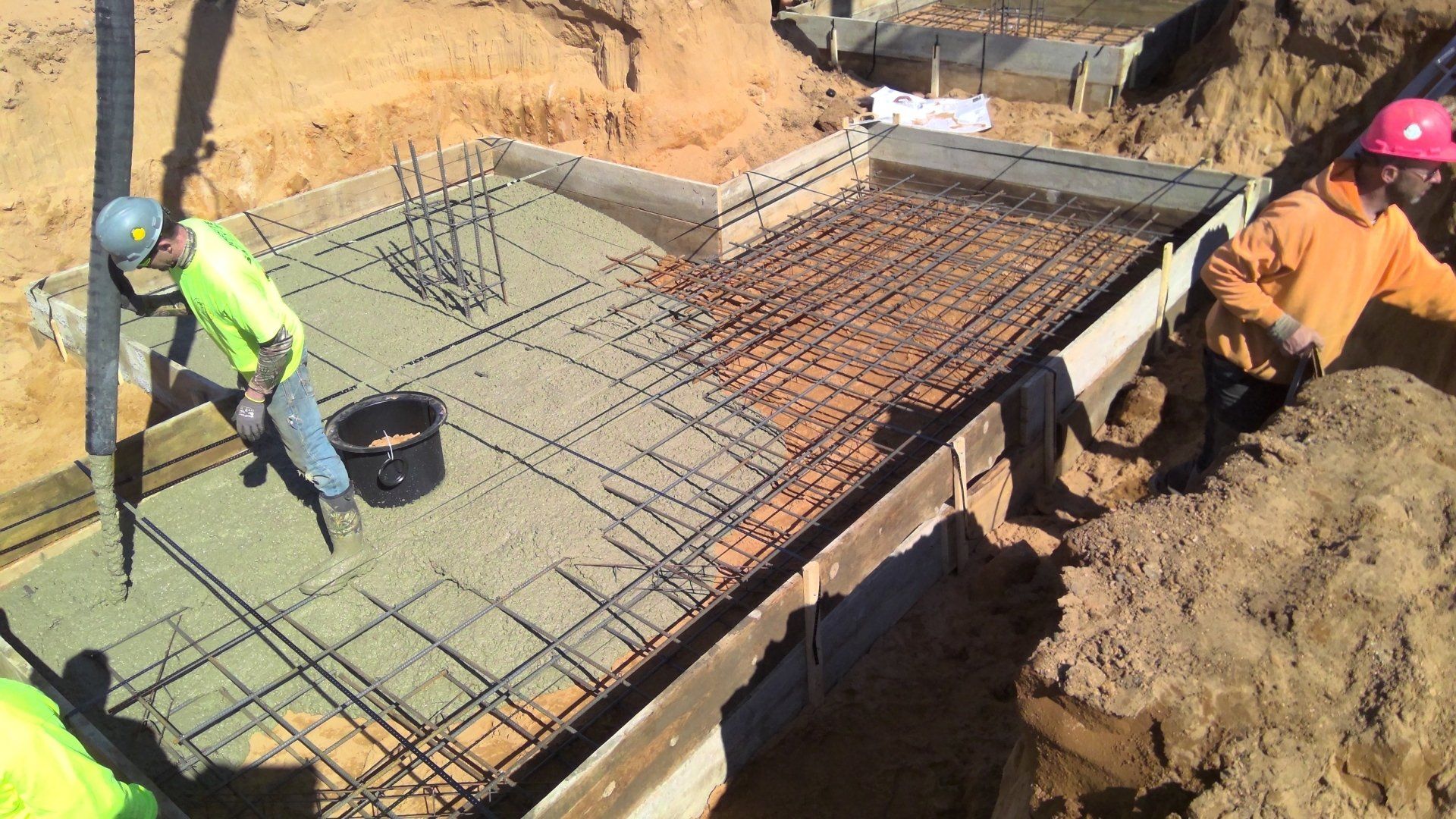 Construction workers pouring concrete on a foundation with rebar. Outdoor setting, sunny day.