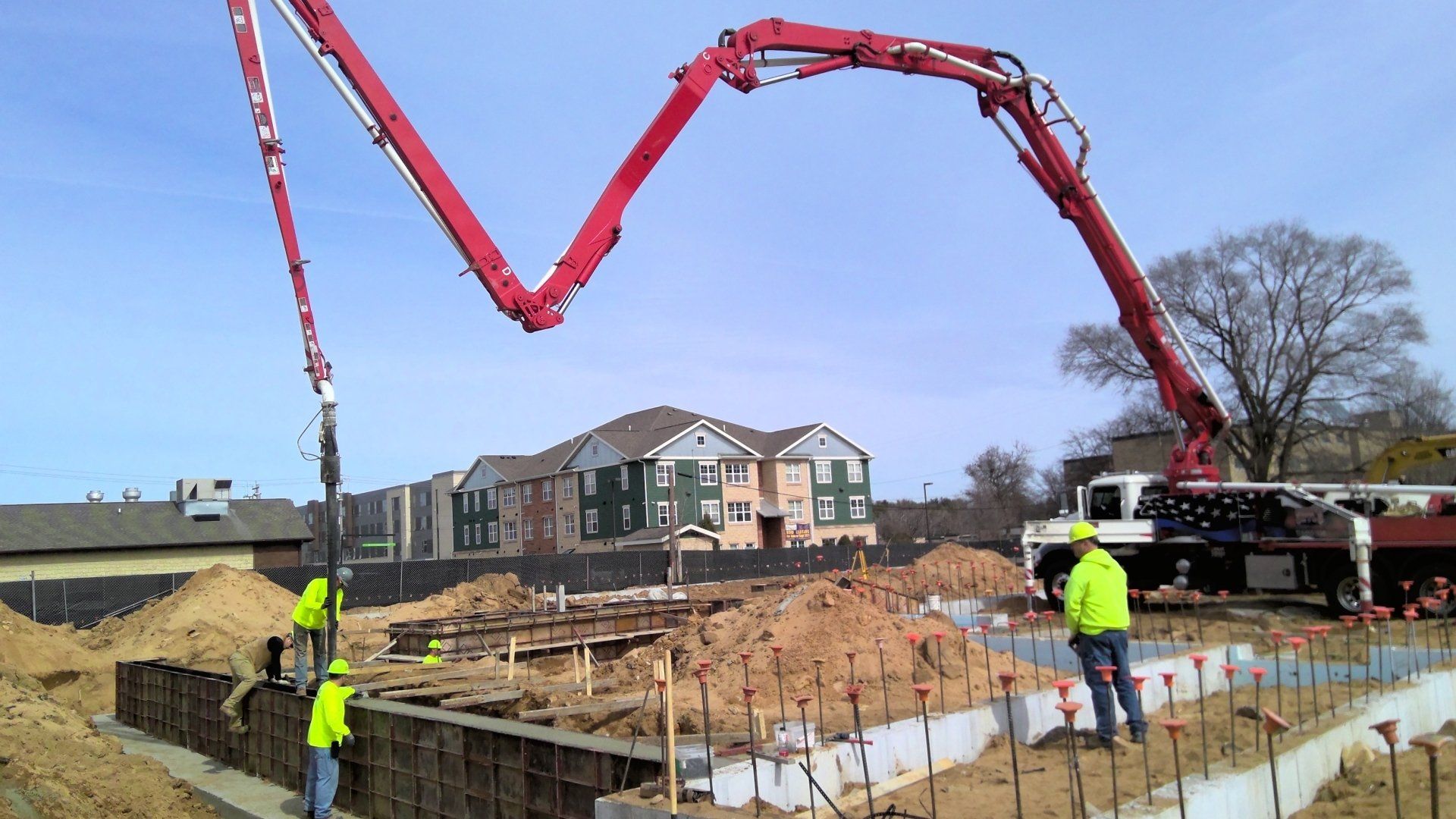 Construction site with concrete pump truck pouring concrete into foundation forms; workers in safety vests.