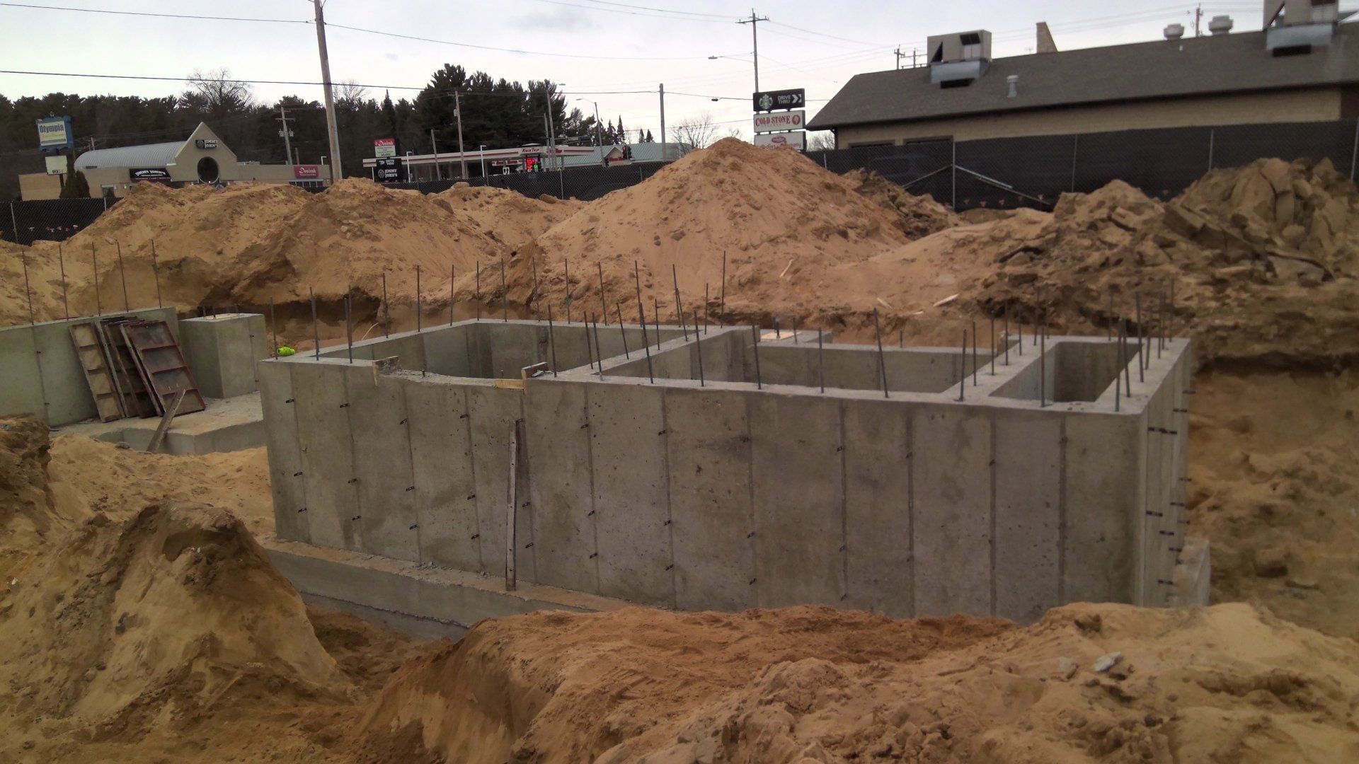 Concrete foundation under construction, surrounded by piles of dirt. Building in the background.
