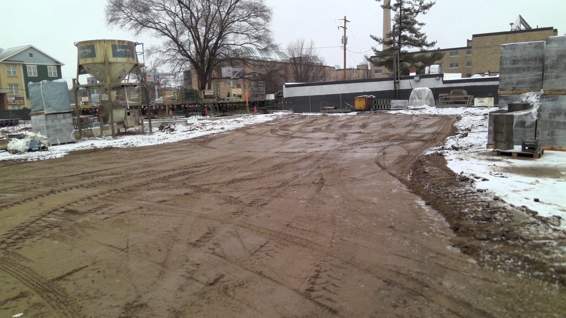Muddy construction site with tire tracks and snow, buildings in the background.