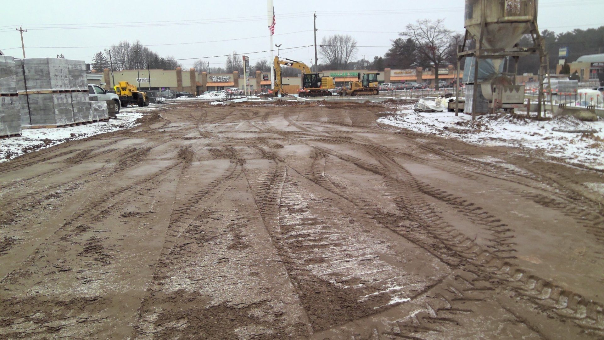 Muddy construction site with tire tracks, heavy machinery, and piles of materials. Snowy ground.