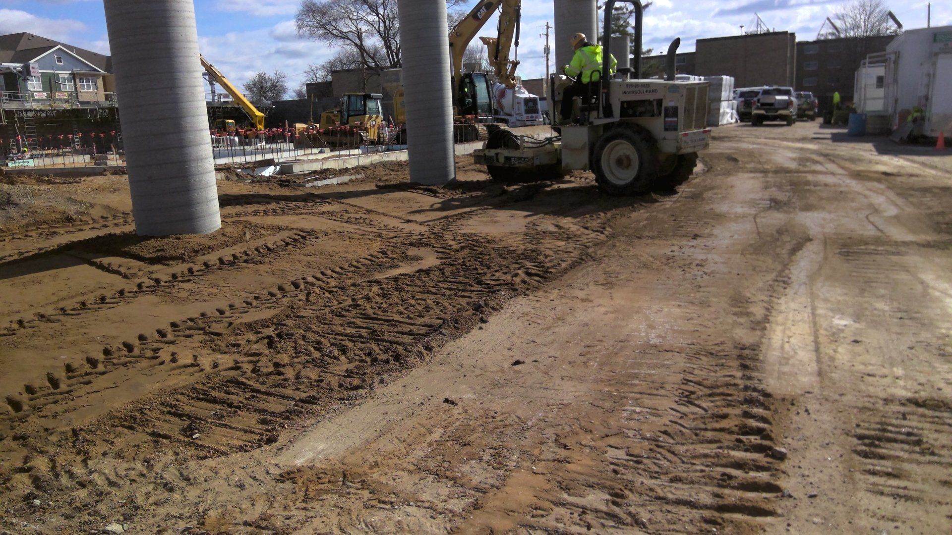 Construction site with large concrete columns, heavy machinery, and dirt road. A worker operates a tractor.