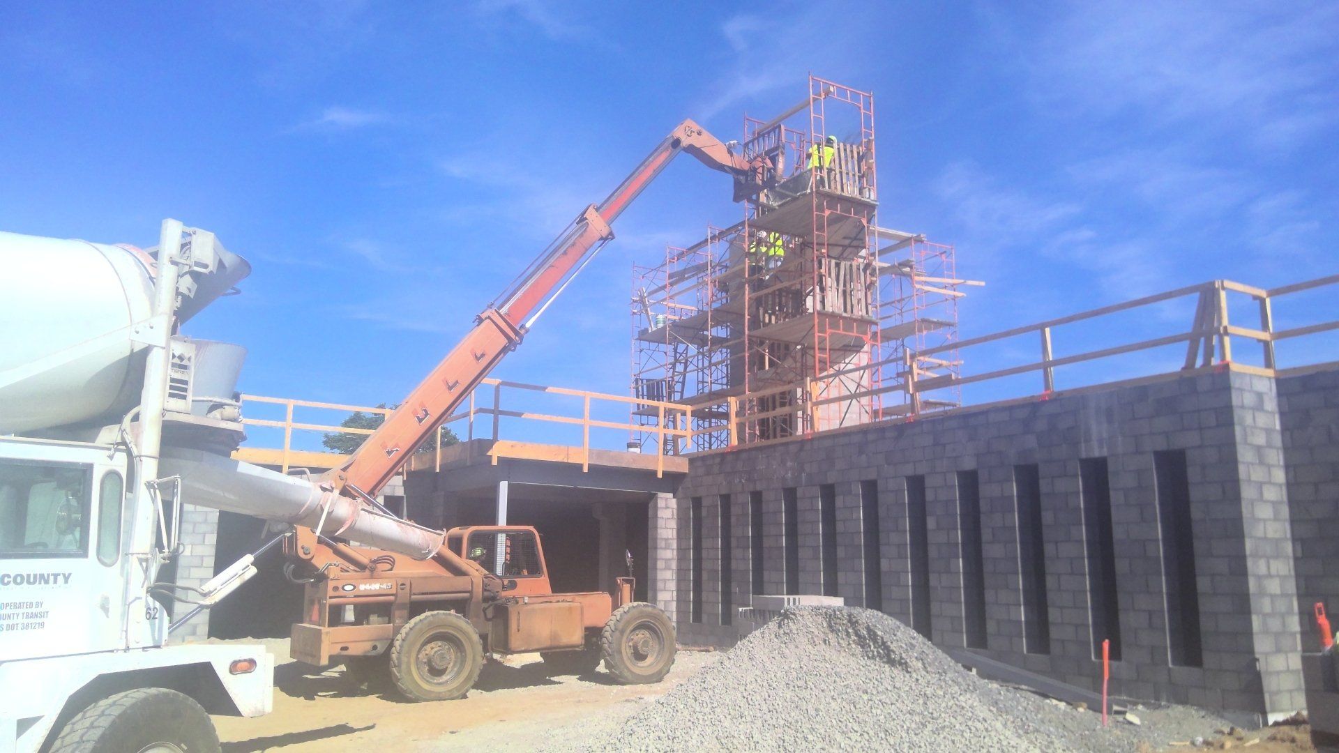 Construction site: Concrete truck pouring concrete near a forklift and scaffolding. Workers are visible on the scaffolding.