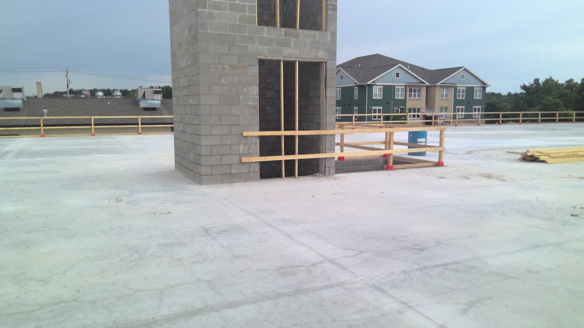 Construction site: concrete structure with temporary wooden safety railing. Apartments and fence in the background.