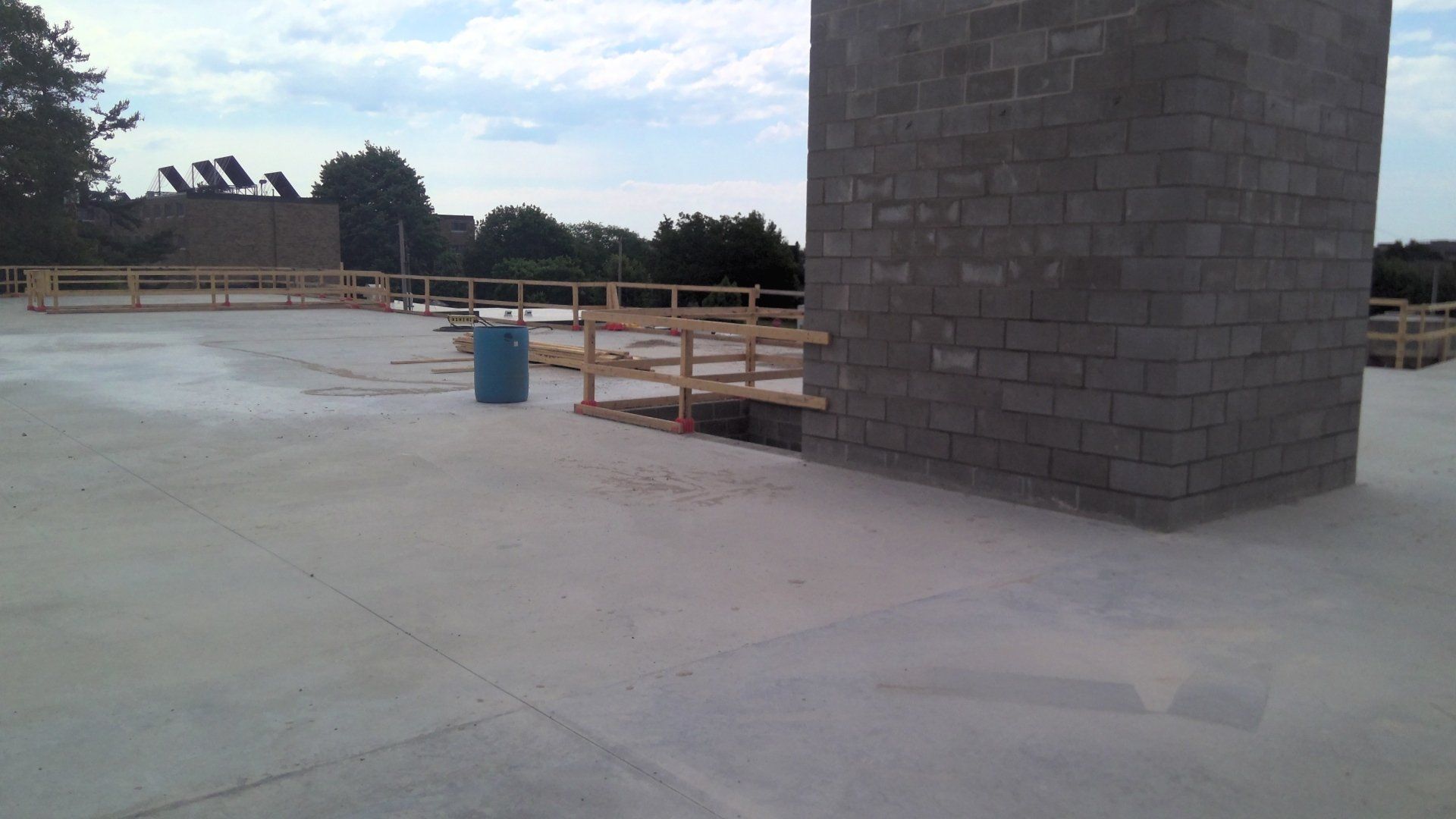Concrete roof with wooden safety railing. Gray block wall, blue trash can, and trees in background.
