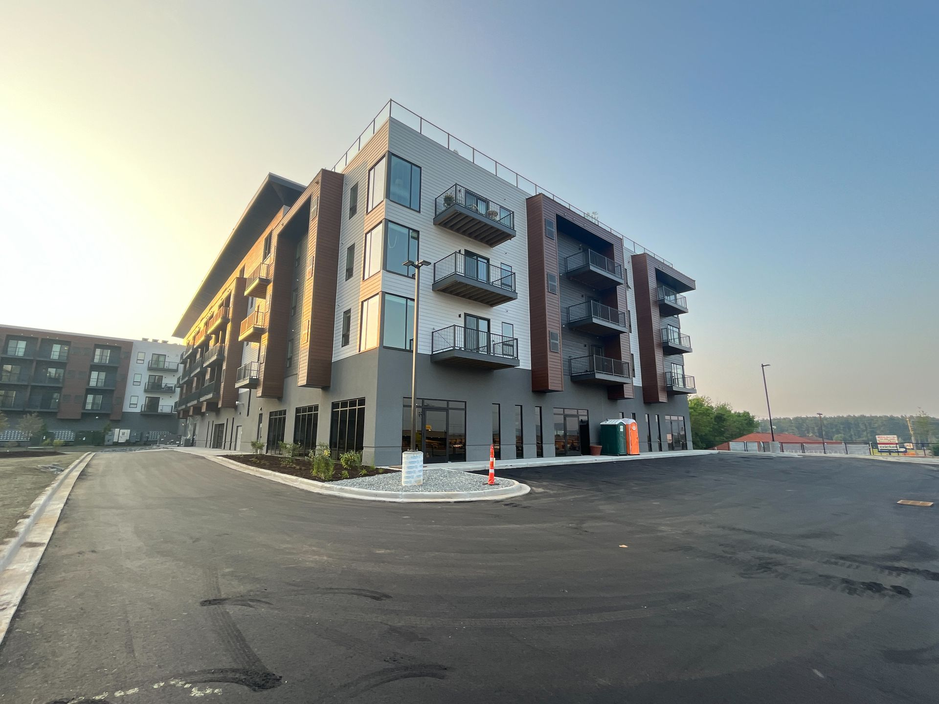 Modern multi-story building with balconies, gray and brown facade, asphalt parking lot.