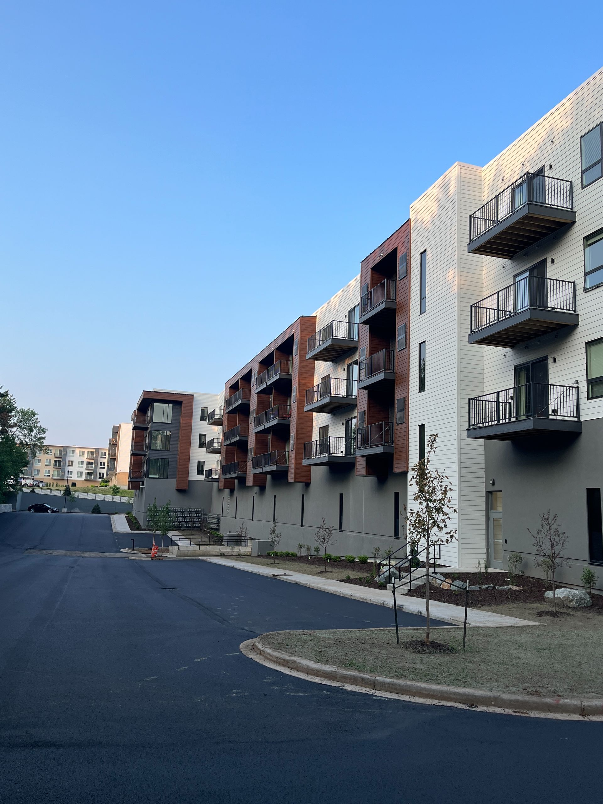 Modern apartment buildings with balconies, against a clear blue sky. A curved road leads to the buildings.