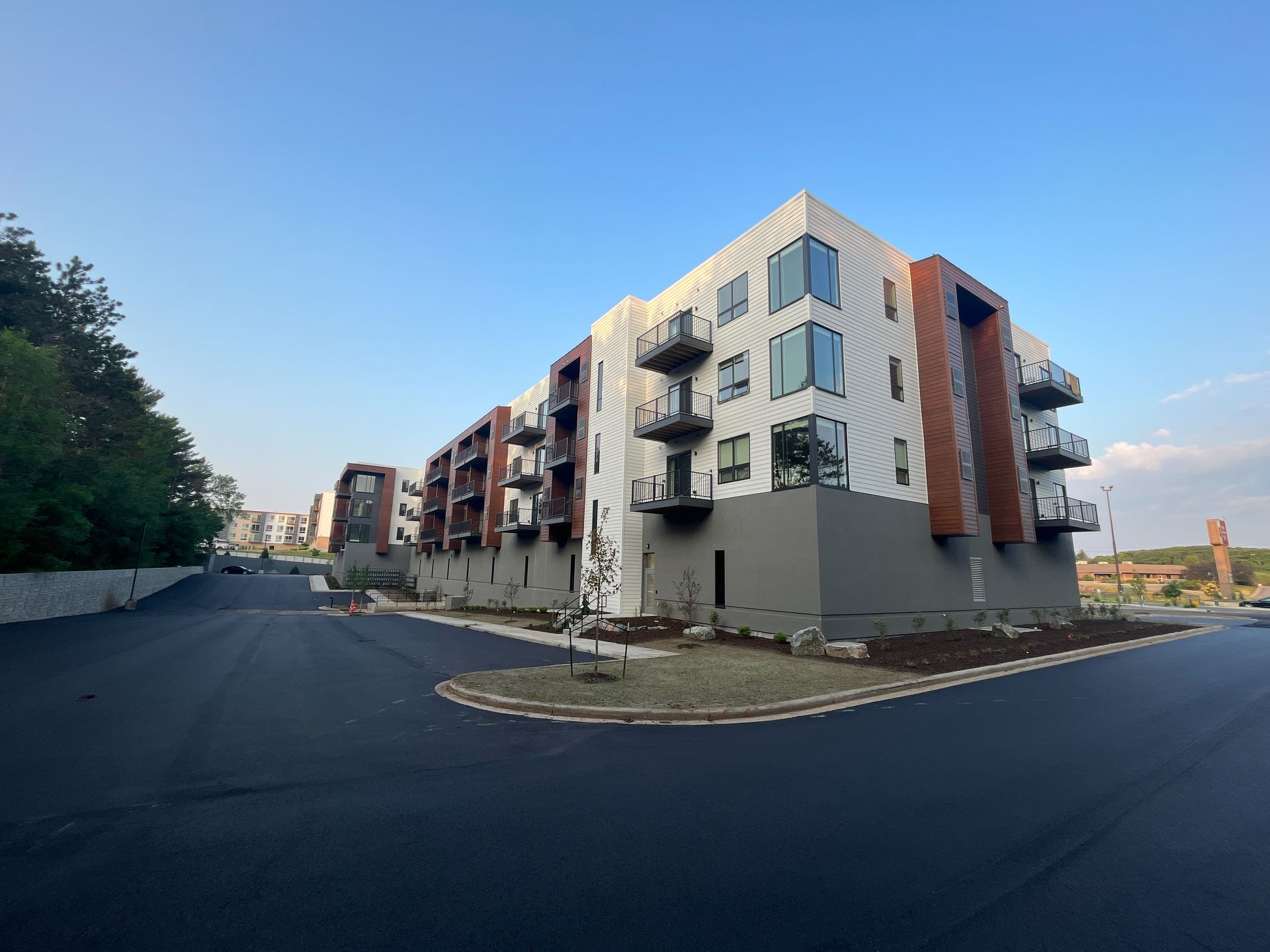 Modern multi-story apartment building with balconies, gray and white siding, and a paved road.