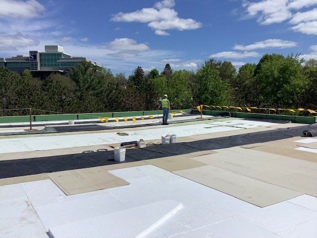 Rooftop construction: A worker stands on a flat roof. White and black materials are spread across the surface, with a building and trees in the background.