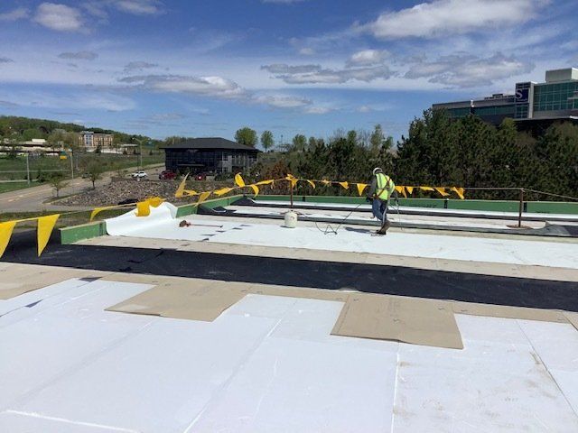 Rooftop construction with a worker, white and black roofing materials, yellow safety flags, and buildings in the background.