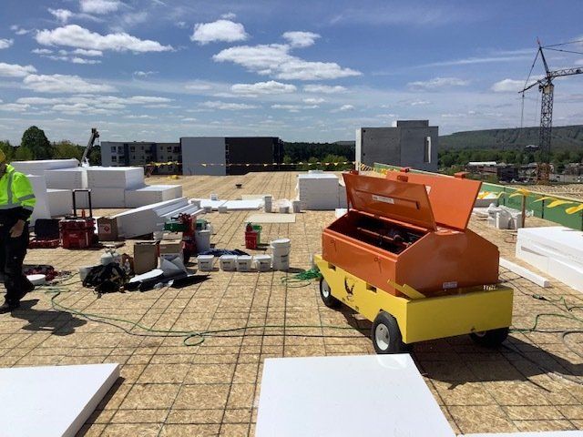 Construction site with orange machinery and white insulation blocks on a rooftop. A person in yellow vest is visible.
