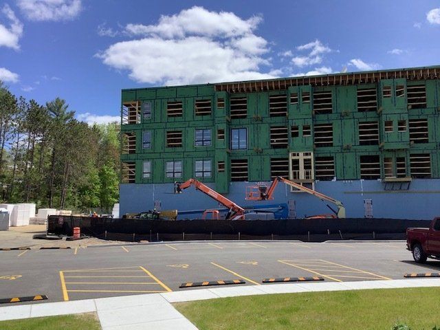 Building under construction, green sheathing, blue tarp, two lifts, black barrier, parked cars, clear sky.