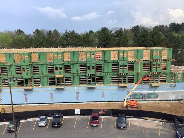 Construction site: three-story building frame with wood siding. Orange lift, cars in a parking lot, trees in background.