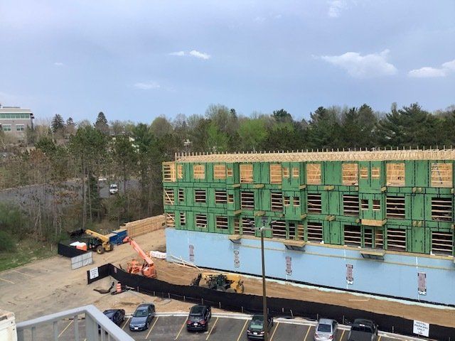 Construction site with a partially built multi-story building. Green sheathing, wood framing, and parked cars are visible.