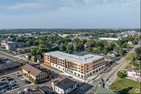 Aerial view of a multi-story apartment building with retail space below. Suburban setting with clear sky.