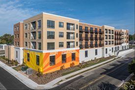 Multi-story apartment building with colorful mural on the lower level, tan and brown facade, blue sky.