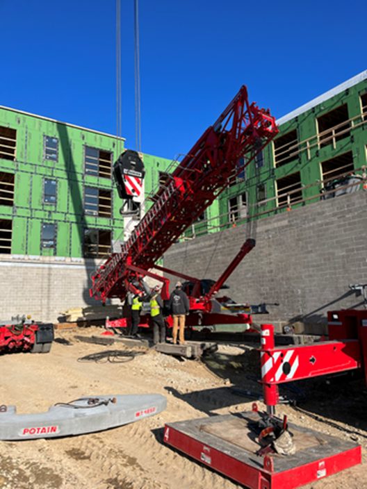 A red crane being assembled at a construction site with workers standing near it and a building in the background.