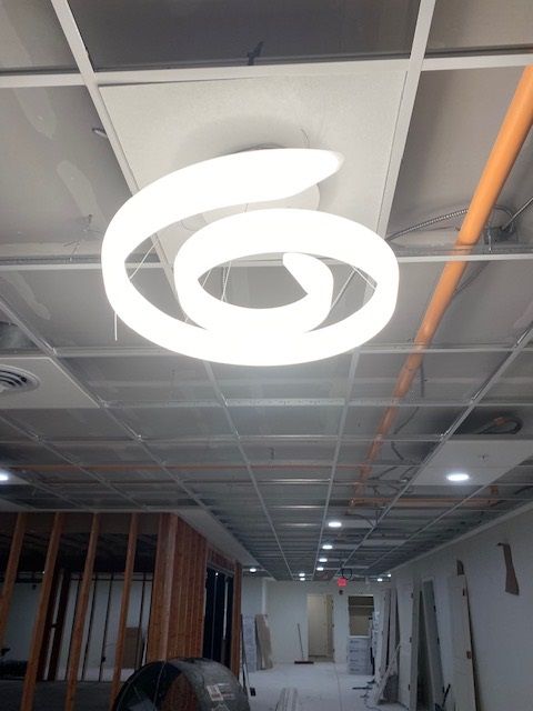 Spiral-shaped, glowing ceiling light fixture in a hallway with exposed ceiling grid and construction materials.