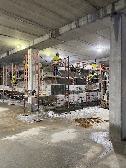 Construction workers on scaffolding inside a building. They wear yellow vests, working on concrete structures.