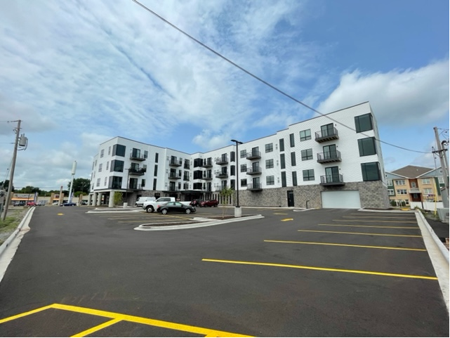 Modern white and gray apartment building with balconies, parking lot in front, and blue sky.