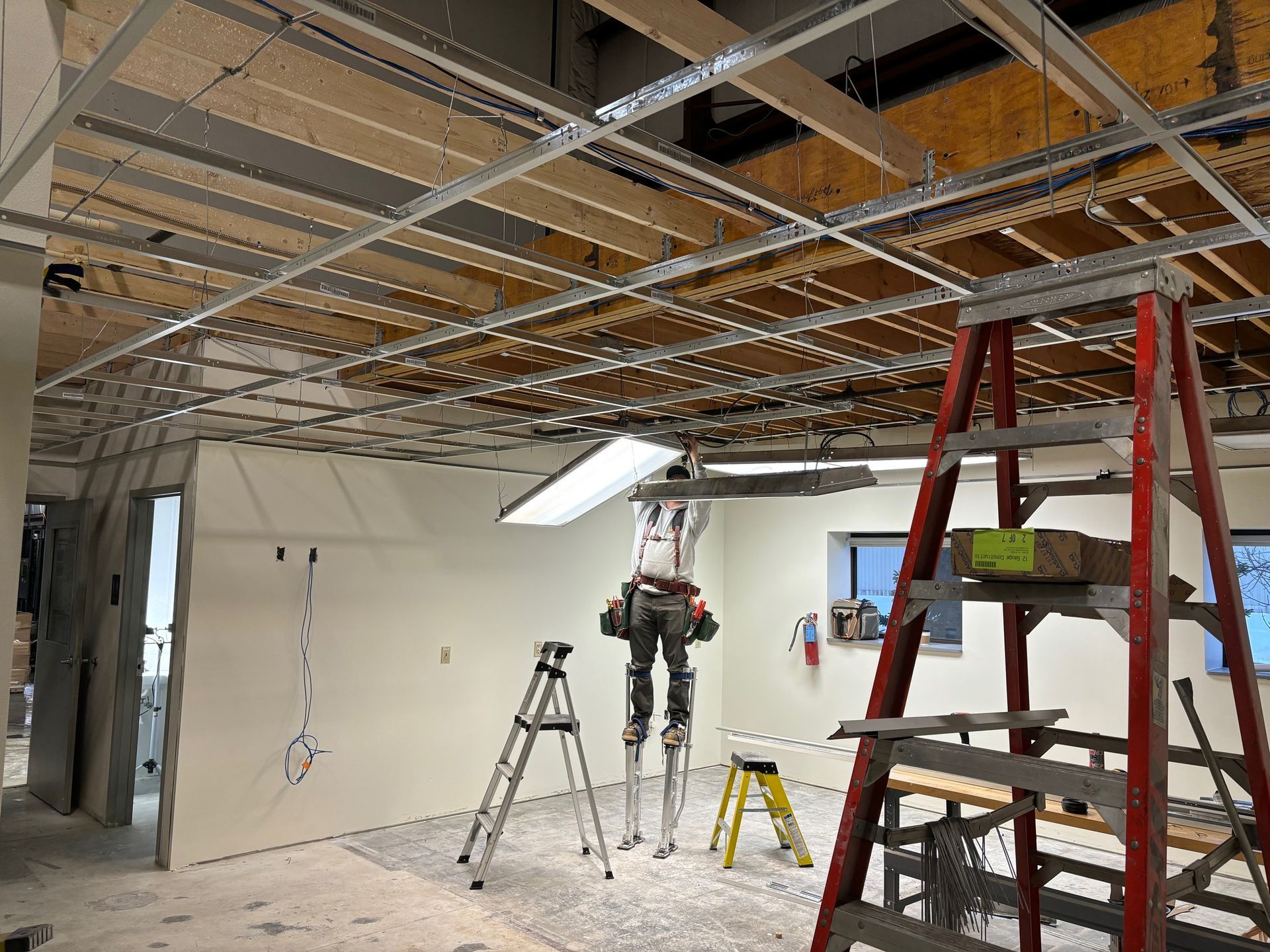 Construction site with exposed ceiling grid, drywall, and ladders. Person on stilts working, tools present.