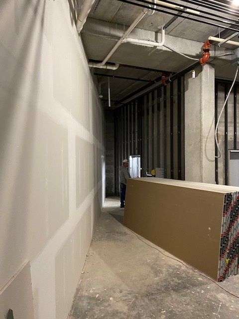 Construction hallway with drywall, exposed pipes, and a person in the distance near a large panel.