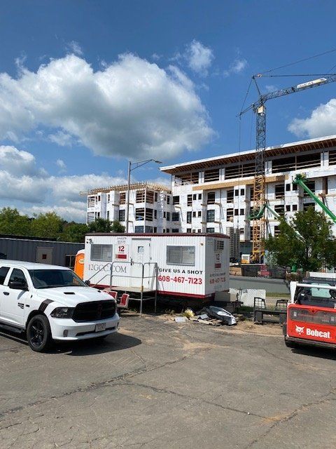 Construction site with partially built buildings, crane, white truck, portable office, and a Bobcat on a sunny day.