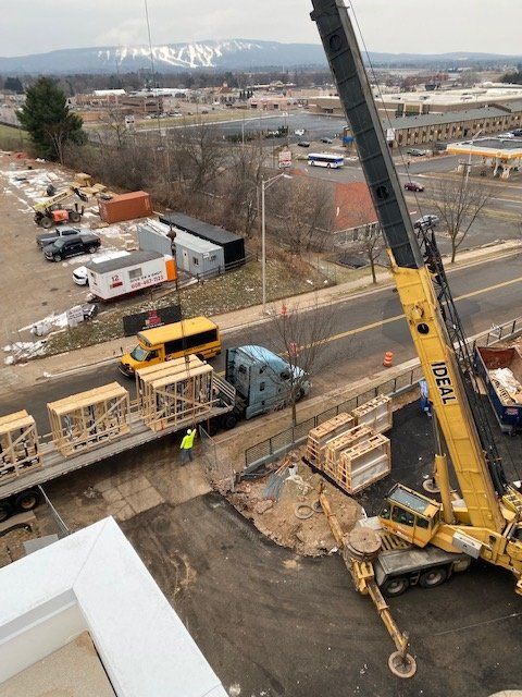 A crane lifts construction materials from a truck beside a building. Yellow crane, workers, and a distant mountain are visible.