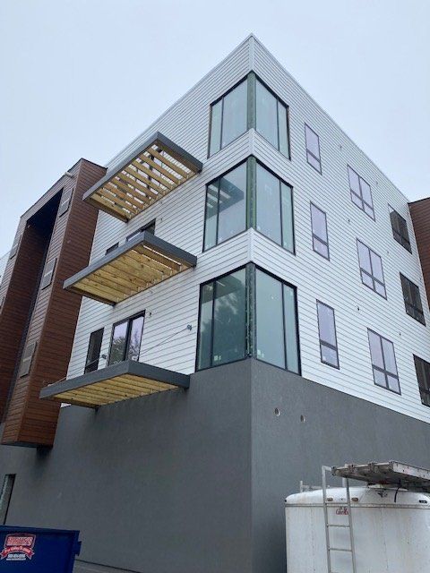 Modern building with white siding, corner windows, and wooden sunshades, grey base, cloudy sky.