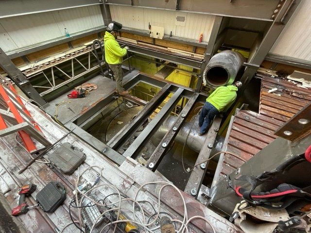 Two workers weld metal beams in a construction site with a large opening. Yellow safety vests.