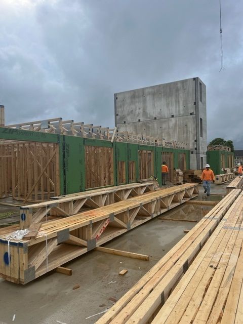 Construction site with wooden trusses and framing; workers in orange vests. Concrete building in background.