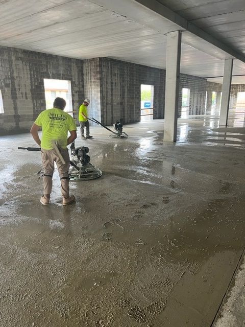 Workers smoothing wet concrete floor with power grinders in a large, open construction space.
