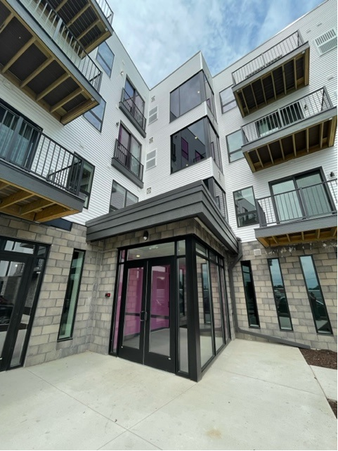 Modern apartment building entrance with glass doors, balconies, and gray and white exterior.