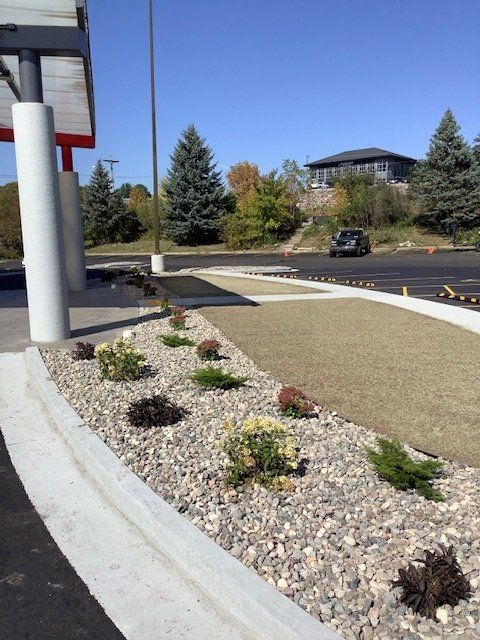 Landscaped area with beige gravel, small plants, and a concrete border, beside a building with white pillars.