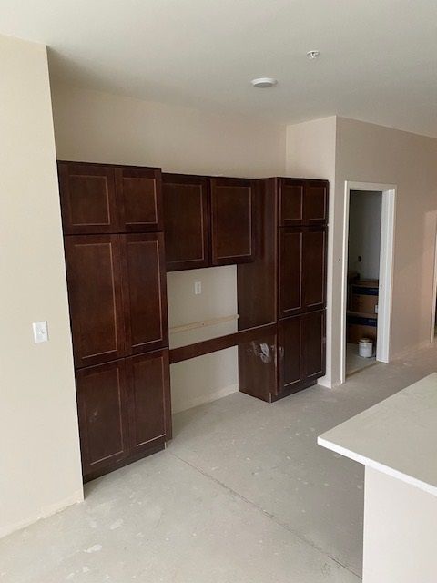 Dark brown kitchen cabinets with a built-in desk, on a light beige wall, in a room under construction.