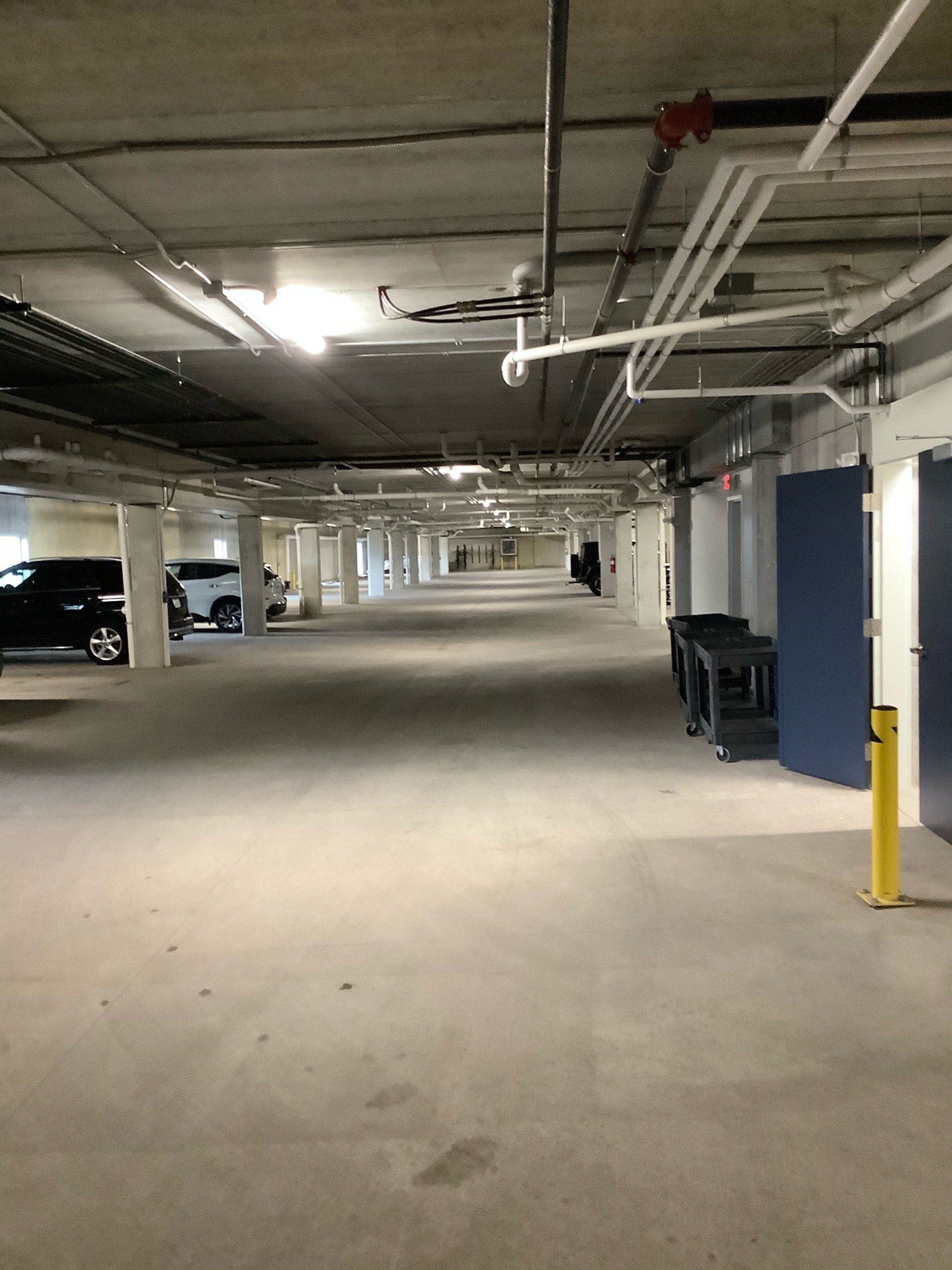 Concrete parking garage with cars, storage units, and overhead pipes.