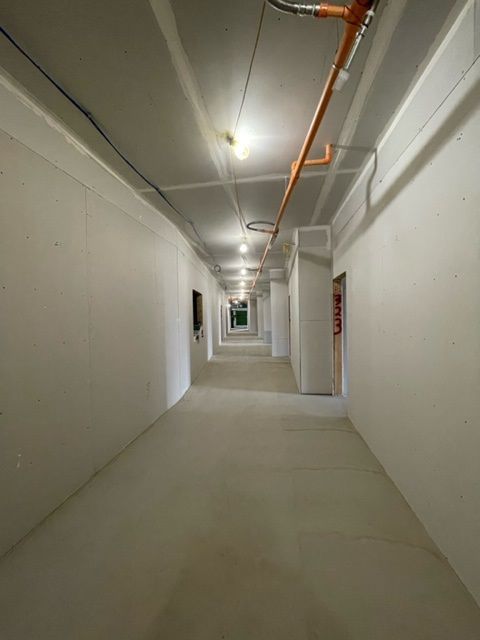Long, unfinished hallway with white walls and ceiling, a row of lights, and a fire sprinkler pipe.