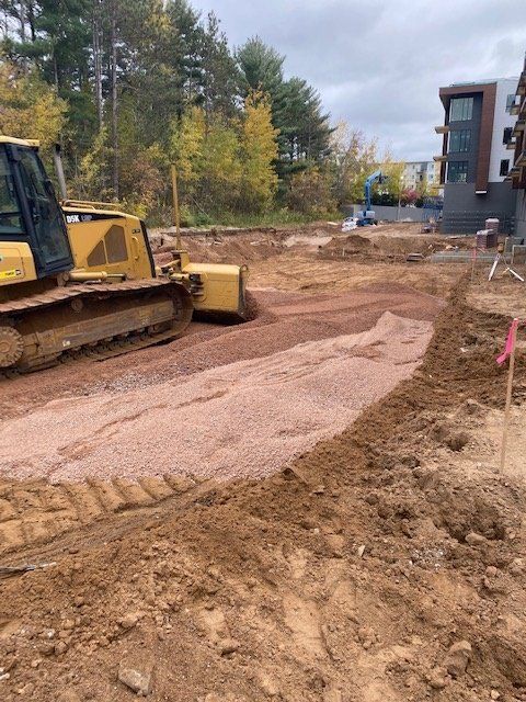 Bulldozer pushing gravel on a construction site. Dirt, trees, and buildings are in the background.