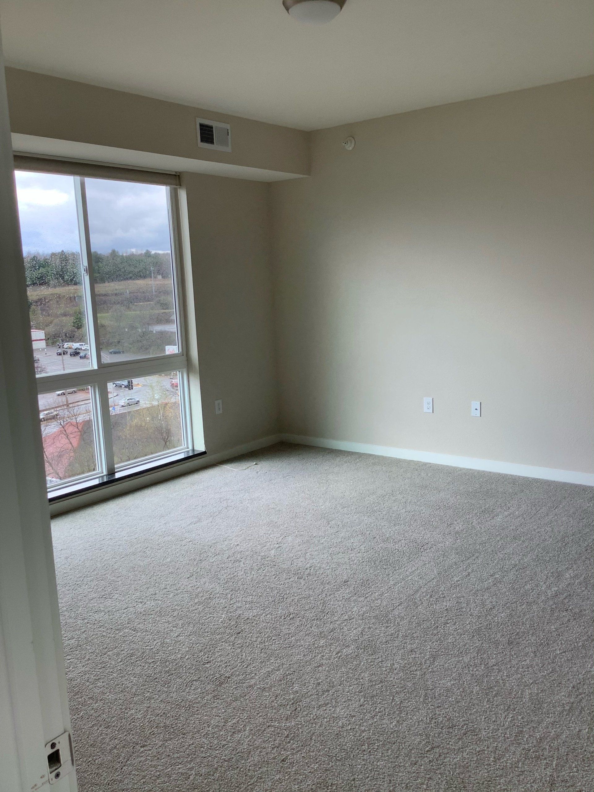 Empty room with large window, beige walls, and gray carpet.