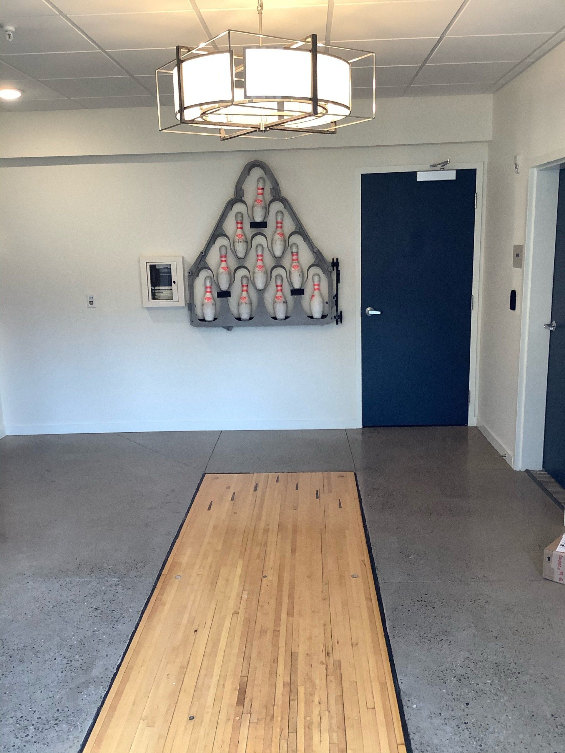 Bowling alley setup: wooden lane, pin display, dark blue door, modern lighting, white wall, gray floor.