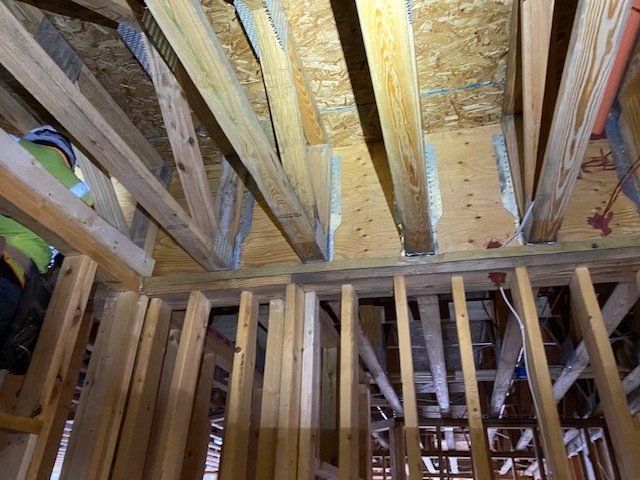 Wooden rafters and studs in a construction setting; exposed structure with plywood sheathing overhead.